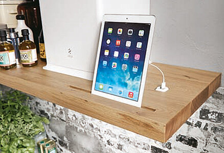 Un cadre de cuisine moderne avec un iPad sur une étagère en bois parmi des bouteilles, connecté à un chargeur, illustrant un mélange de technologie et de confort domestique.