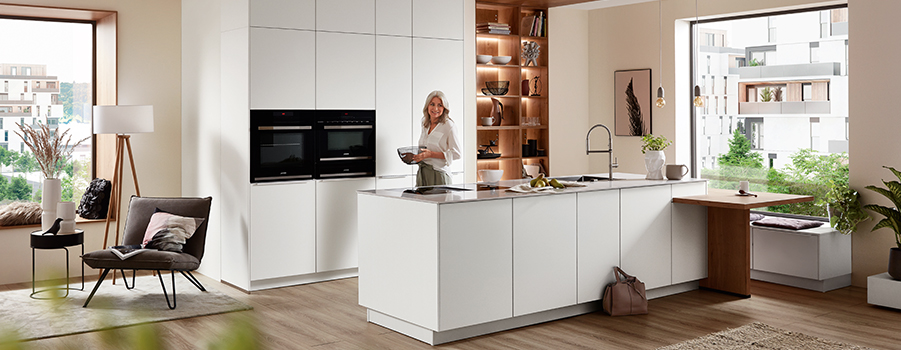 Diseño de cocina moderna con gabinetes blancos limpios, una isla central, y una persona cocinando en un espacio luminoso y aireado con vista a la ciudad a través de grandes ventanas.