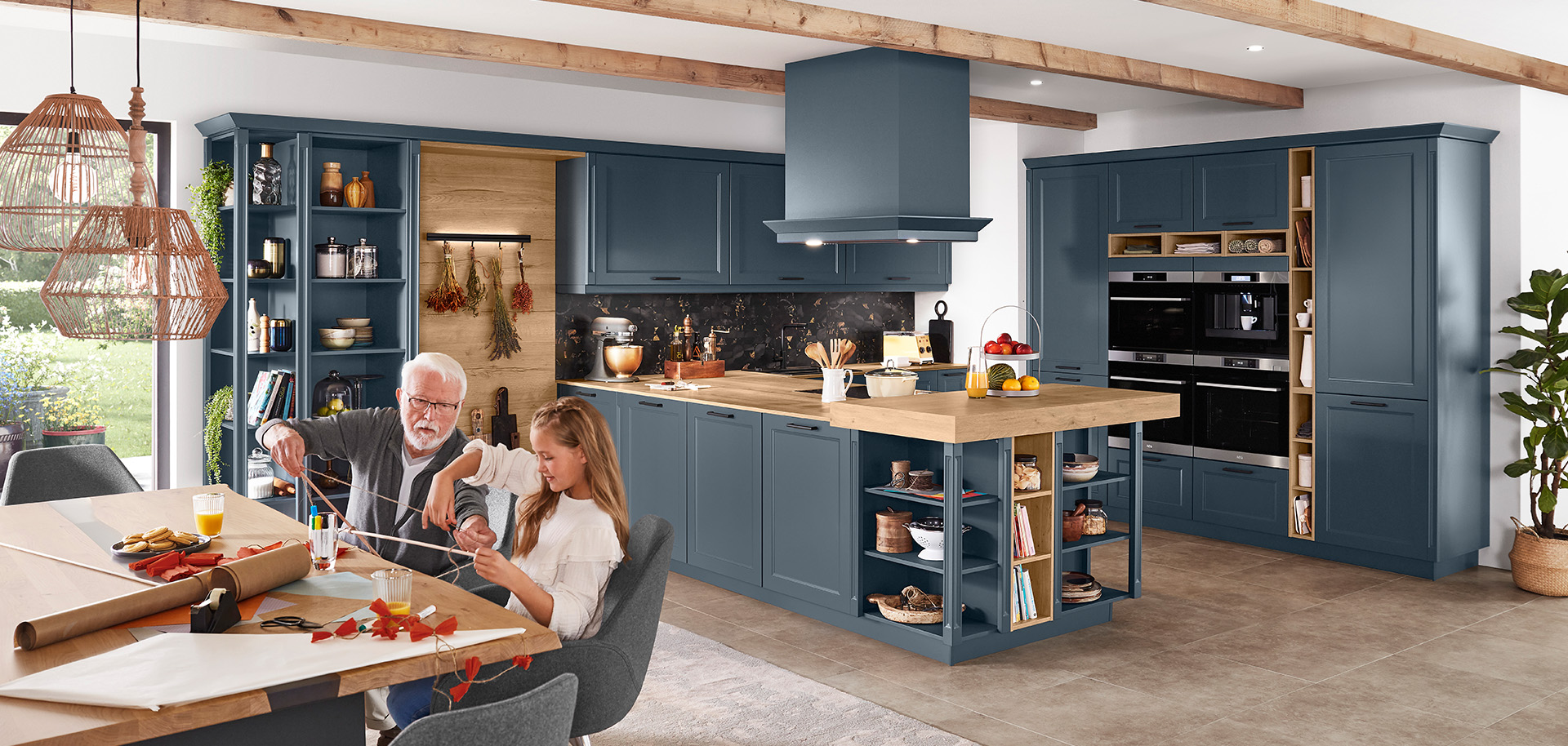 Een ruime, moderne keuken met marineblauwe kasten, roestvrijstalen apparaten en een oudere persoon die met een kind aan een zonovergoten houten tafel knutselt.