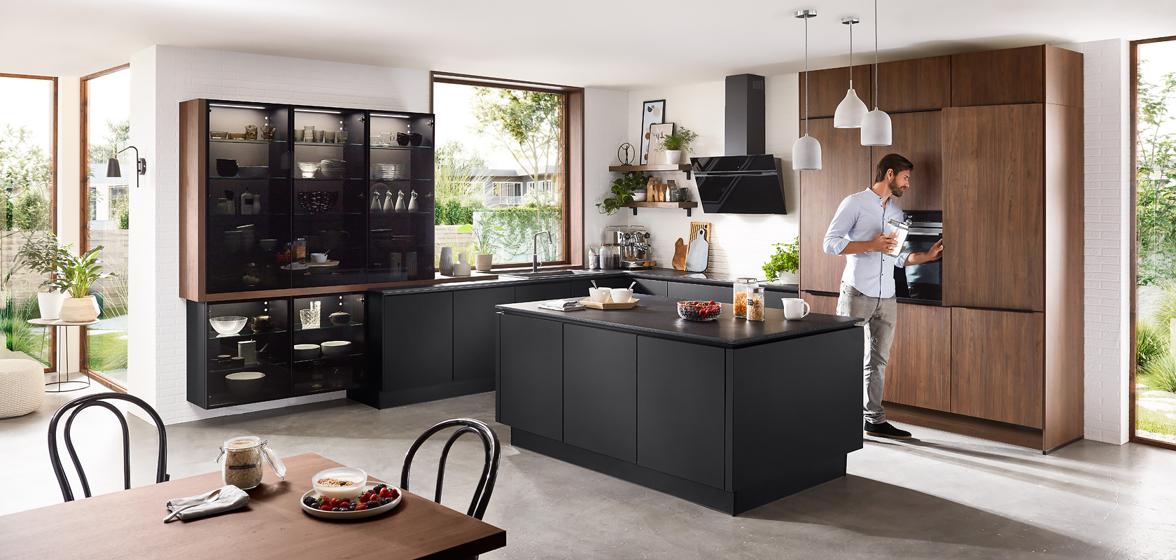 Intérieur de cuisine moderne avec des armoires noires élégantes, des accents en bois, et un homme préparant une boisson sur le comptoir, offrant une ambiance à la fois chaleureuse et contemporaine.