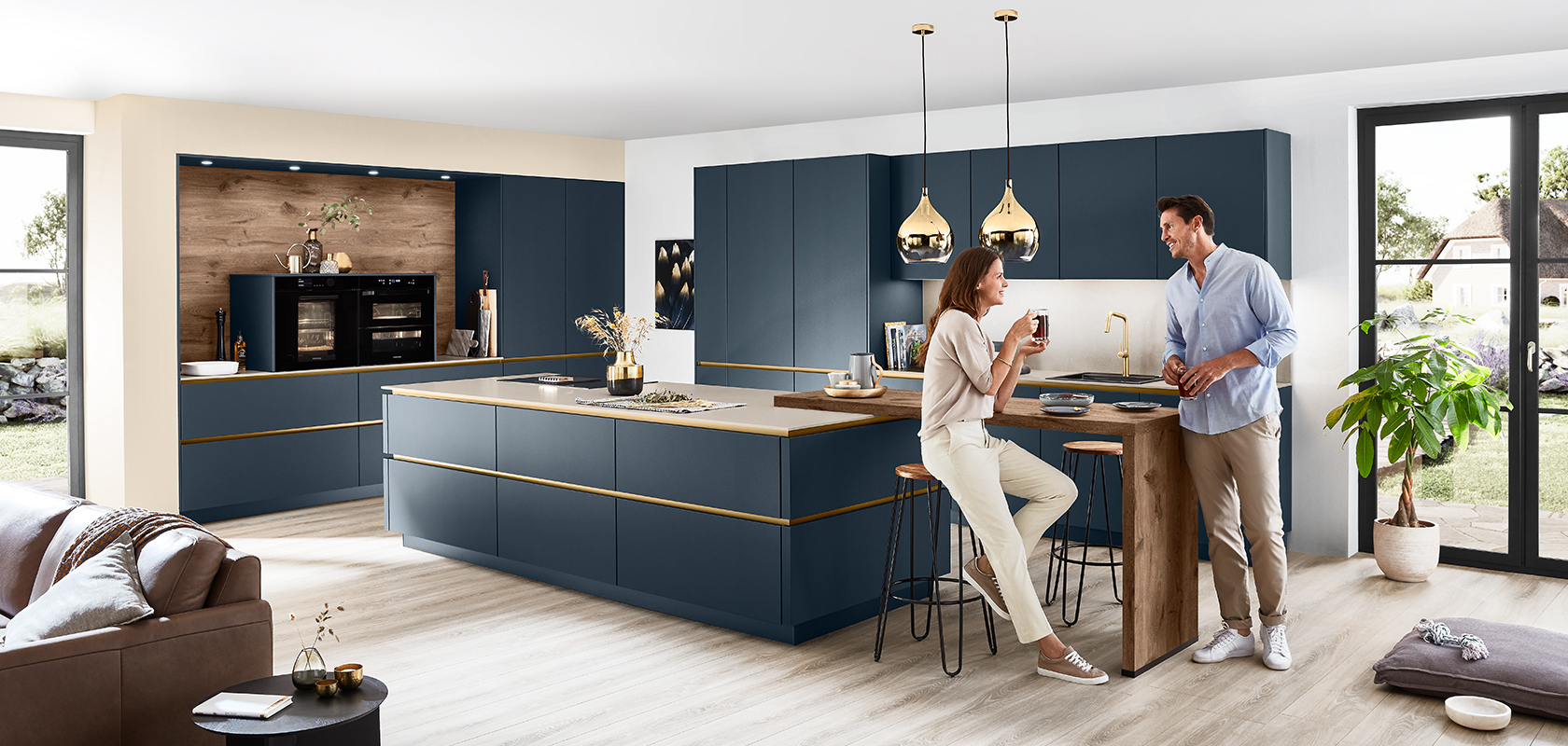 Interior de cocina moderna con una pareja disfrutando de vino alrededor de una isla elegante con gabinetes de color azul marino y elegantes detalles dorados, bajo cálidas luces colgantes.