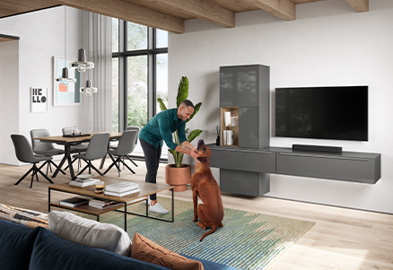 Un hombre en una sala de estar moderna juega con un perro marrón animado, rodeado de muebles elegantes, acentos de madera y una estética de diseño contemporáneo y acogedor.