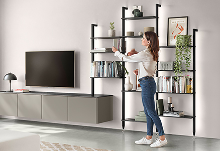 Soggiorno moderno con un'elegante unità di scaffalature montata a parete con una donna che dispone libri, adiacente a una TV a schermo piatto e mobili minimalisti.