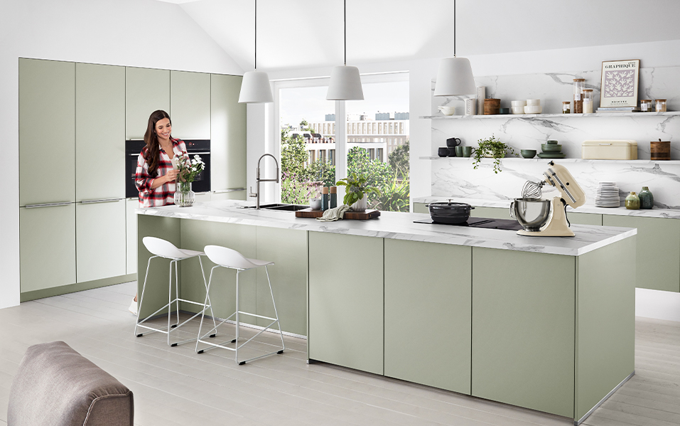 A modern kitchen featuring sleek green cabinetry, marble countertops, and stainless-steel appliances, with a person enjoying the elegant, well-lit space.