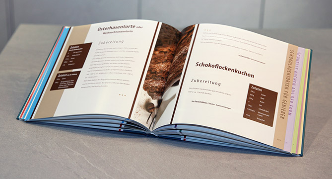 Abre el libro de cocina en una encimera de cocina mostrando recetas, resaltando la página de "Schokoflockenkuchen" con instrucciones de preparación visibles.