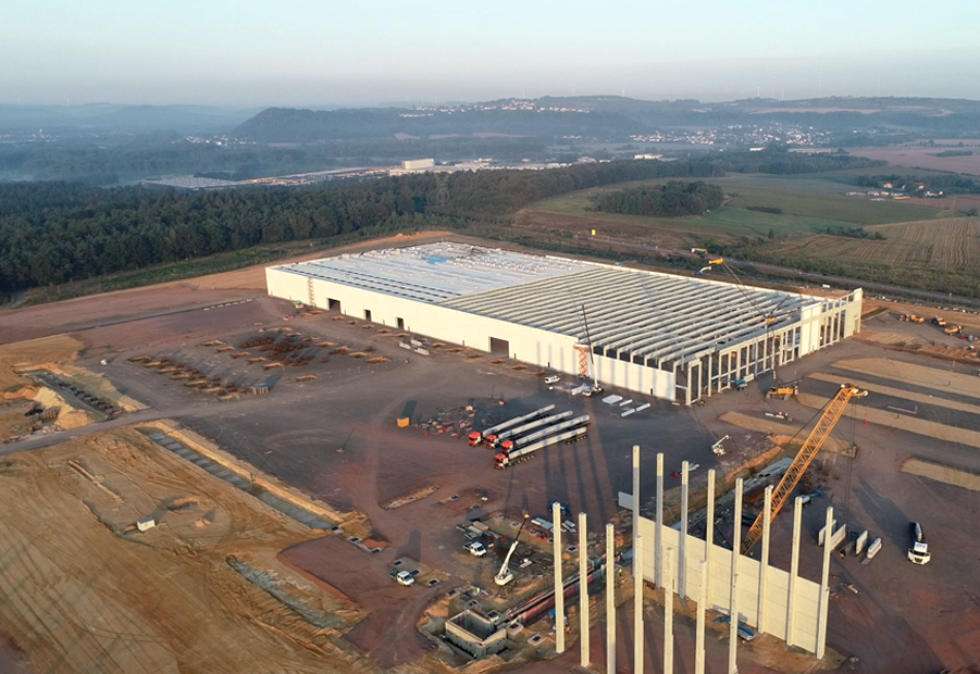 Vista aérea de un gran sitio de construcción industrial con una estructura de almacén parcialmente terminada, rodeada de vehículos de construcción y equipo en terreno abierto.