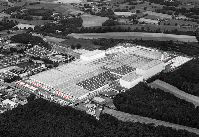Luchtfoto in zwart-wit van een industrieel complex met grote fabrieksgebouwen omringd door groene velden en een paar clusters van kleinere structuren.