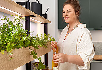 Mujer en una cocina moderna cuidando su jardín de hierbas interior, bañado por luz natural, mostrando un estilo de vida saludable y sostenible en casa.