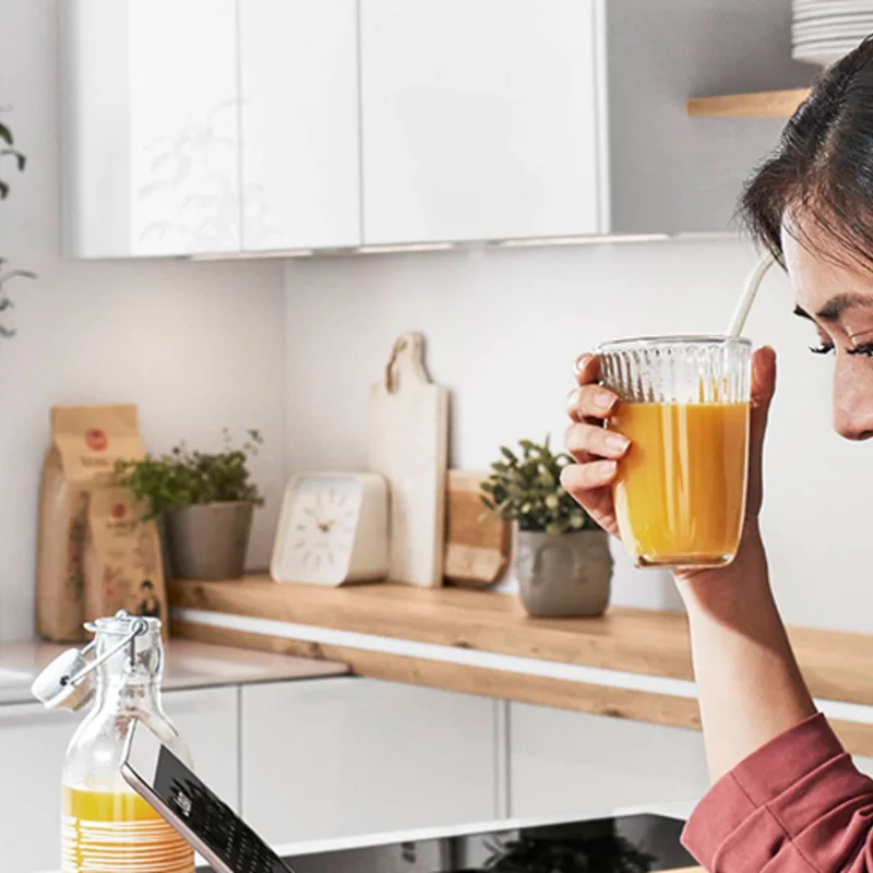 Moderne Küche mit Front NOVALUX 516 in Alpinweiß Hochglanz, warme Holzarbeitsplatte, wohnliche Details und eine Frau, die entspannt ein Glas Orangensaft genießt.