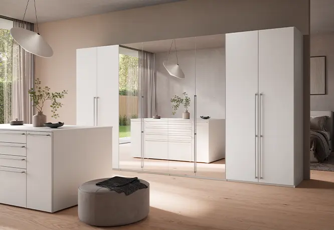 Schlafzimmer mit weißen Kleiderschränken, Spiegelfront, Kommode und rundem Sitzhocker in heller Raumgestaltung.
