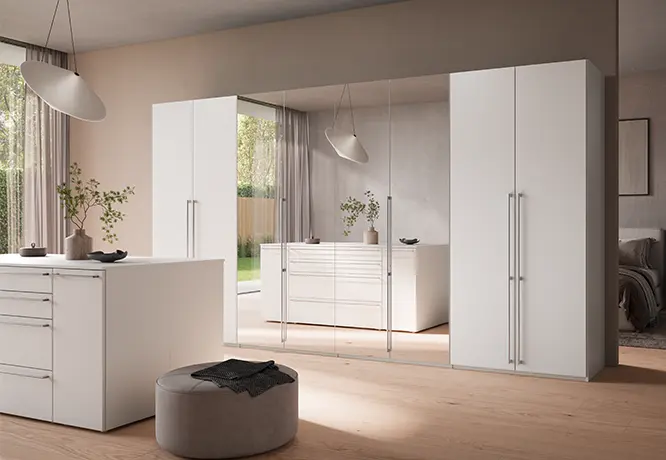 Schlafzimmer mit weißen Kleiderschränken, Spiegelfront, Kommode und rundem Sitzhocker in heller Raumgestaltung.