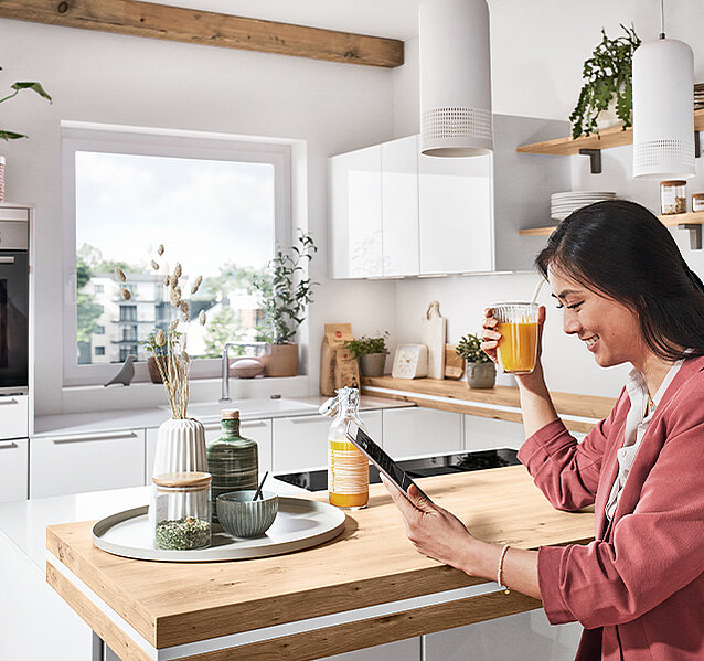 Žena si užívá své ranní čaje při prohlížení tabletu v moderní, světlé kuchyni s stylovými dřevěnými prvky a zelenými rostlinami.