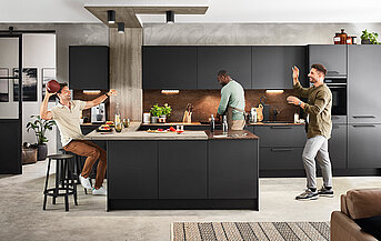 Tři muži si užívají neformální, zábavné setkání v moderní kuchyni s elegantními černými skříňkami, připravují jídlo a sdílí smích.