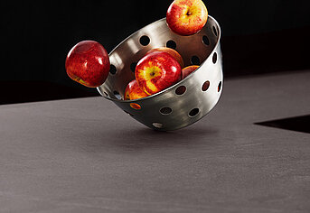 A stainless steel colander tips, spilling ripe red apples onto a dark contrasting surface, showcasing a blend of culinary utility and fresh produce.