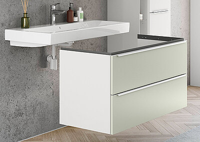 Vanity de baño moderno con líneas limpias que presenta un lavabo blanco, cajones verdes salvia y blancos, en contraste con una pared gris texturizada.