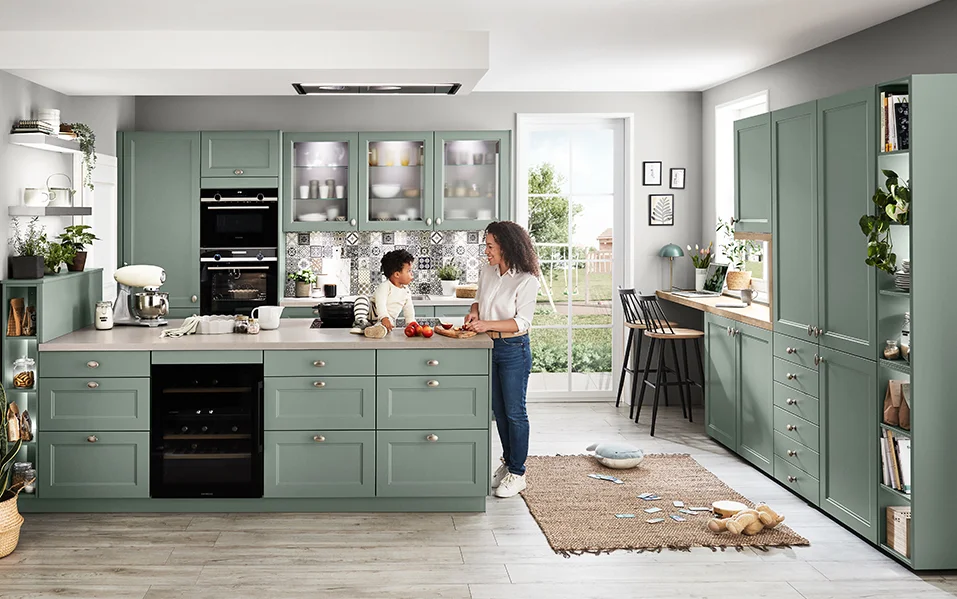 Przytulna wiejska kuchnia z frontem CASCADA 776 w kolorze trzcinowej zieleni, wyspą kuchenną, zintegrowanym domowym biurem i żywą rodzinną atmosferą.