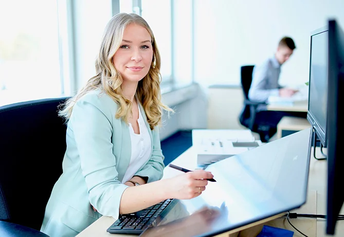 Eine professionelle Frau in einem hellblauen Blazer arbeitet an einem Schreibtisch mit einem Computer in einer hellen, modernen Büroumgebung.