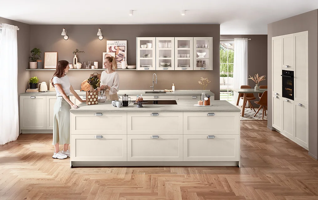 Cucina moderna in stile country in sabbia opaca con frontale Nordic 788, isola di cottura, linee chiare e piano di lavoro in travertino chiaro su una parete dal tono caldo.