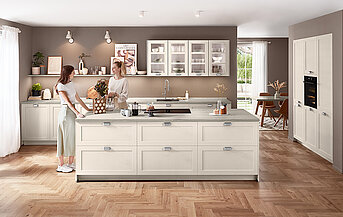 Moderne Landhausküche in Sand matt mit Front Nordic 788, Kochinsel, klarer Linienführung und heller Travertin-Arbeitsplatte vor warmem Wandton.