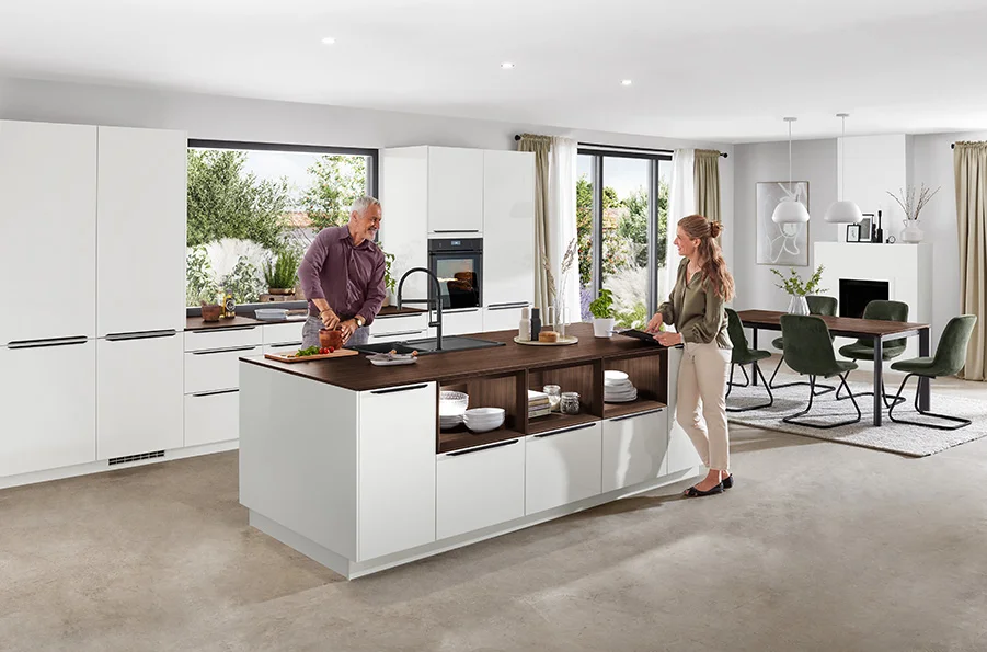 Cucina spaziosa con frontale NOVALUX 512 in bianco lucido, isola di cottura con soluzione a scaffale aperto e piano di lavoro in legno scuro, due persone che cucinano insieme in un soggiorno moderno e luminoso.