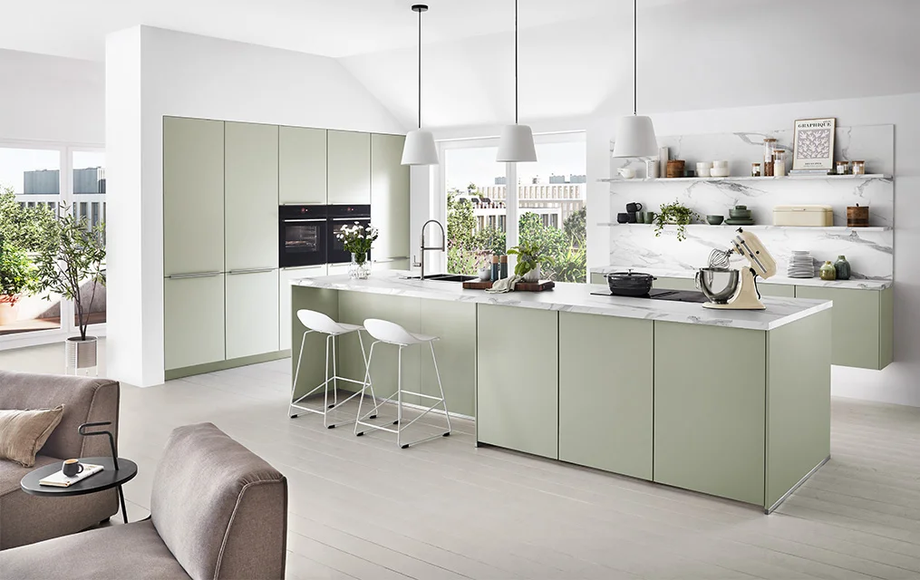 Cucina senza maniglie di colore chiaro con frontale SENSO 494 in Jade Premium opaco, isola di cottura con piano di lavoro in marmo e ripiani aperti davanti a un'ampia finestra frontale nel soggiorno moderno.