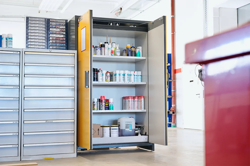 Un taller bien organizado con un armario de almacenamiento alto y abierto que muestra varias latas, botellas y equipos, junto a una unidad de cajones de metal.