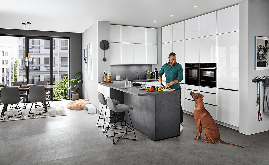 Nowoczesne wnętrze kuchni z mężczyzną przygotowującym jedzenie na blacie, podczas gdy pies siedzi obok, prezentujące eleganckie urządzenia AGD i dobrze oświetloną jadalnię.