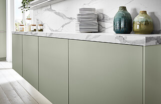 Moderní kuchyně s čistými liniemi, která má šedozelené skříňky, mramorové pracovní desky a minimalistické uspořádání keramických nádob a váz.