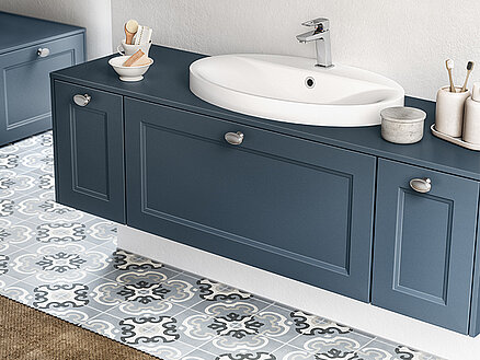 Elegant bathroom interior featuring a modern navy vanity with a white basin, silver faucet, and tasteful decorative accessories on a patterned tile floor.