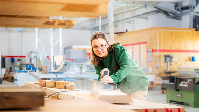Auszubildende in der Tischlerei bei der Produktion, während sie ein Holzstück mit einer Hobelmaschine bearbeitet – handwerkliche Ausbildung mit Präzision.