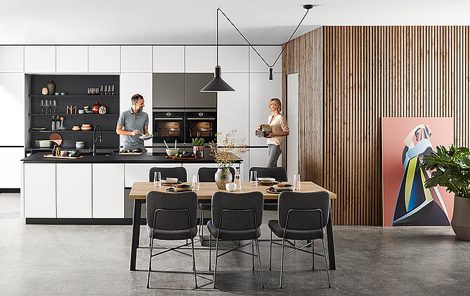 Una cocina moderna con una pareja cocinando y charlando, con gabinetes elegantes en blanco y negro, acentos de madera y un área de comedor con muebles elegantes.