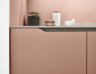 Współczesny design kuchni z eleganckim blatem i minimalistyczną dekoracją na tle delikatnego różowego koloru, łączący funkcjonalność z nowoczesną estetyką.