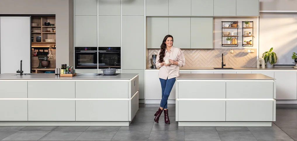 Sally steht in einer großzügigen Küche mit zwei Kochinseln, offenen Regalelementen und harmonischer Lichtstimmung. Das Bild zeigt modernes Küchendesign mit viel Raum für Kochen, Backen und Wohnen.