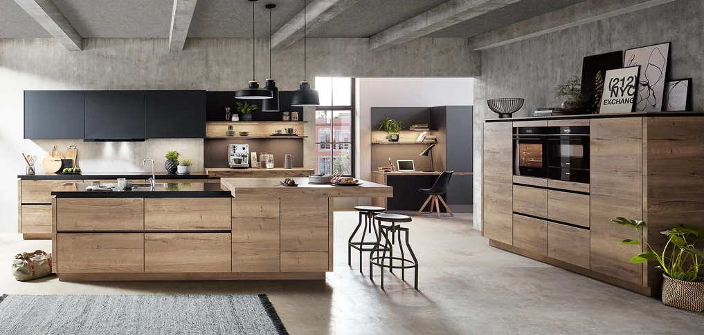 Cucina open space con frontale STRUCTURA 402 in rovere avana, isola di cottura moderna con effetto legno e accenti neri, area di lavoro adiacente con soluzione scrivania, stile loft urbano con pareti in cemento a vista.