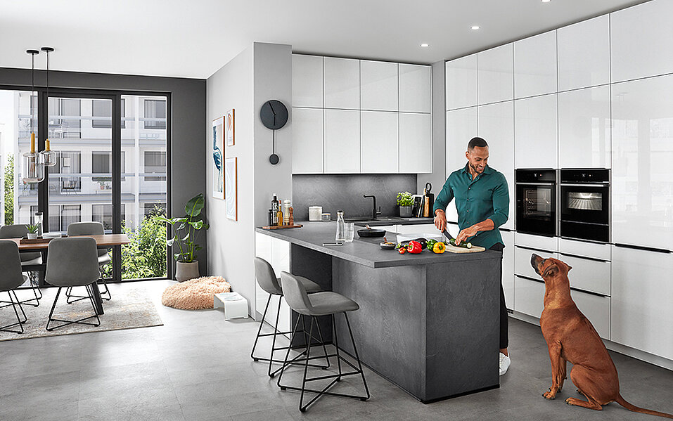 Intérieur de cuisine moderne avec un homme préparant de la nourriture sur le plan de travail pendant qu'un chien brun se tient à proximité, le regardant dans une pièce bien éclairée et spacieuse.