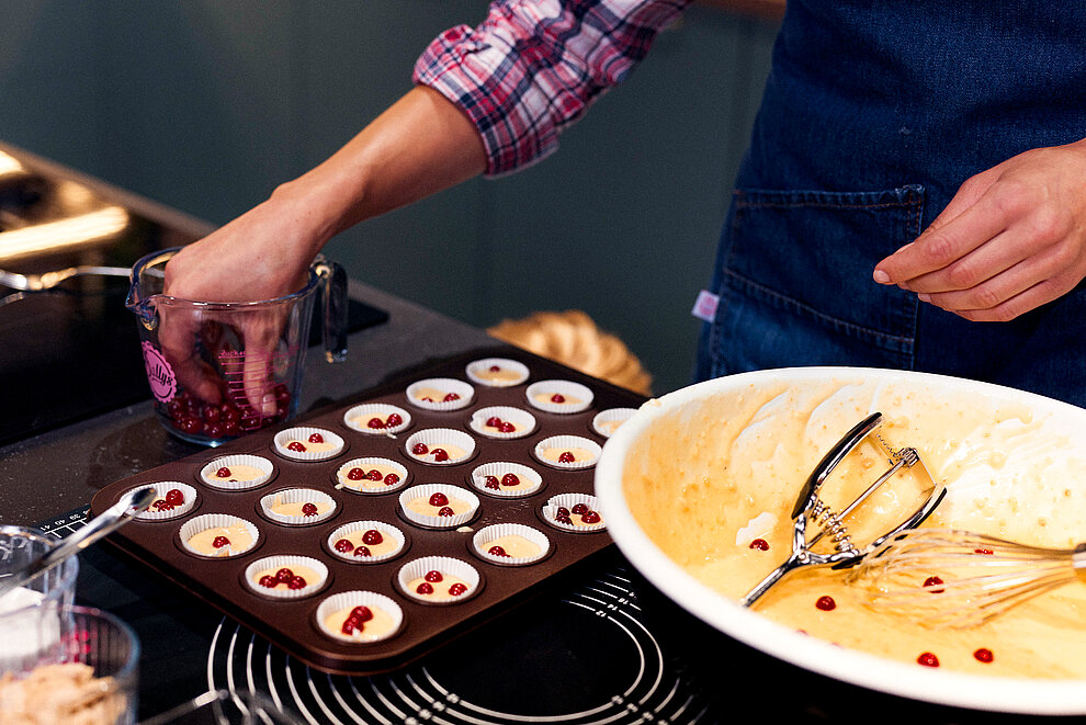 A person in a plaid shirt and denim apron prepares raspberry cupcakes, placing fresh berries atop batter in a muffin tin.