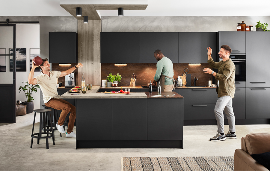 Trois individus apprécient un cadre de cuisine moderne, avec deux hommes cuisinant joyeusement et un assis à un comptoir interagissant de manière décontractée avec une tartine.