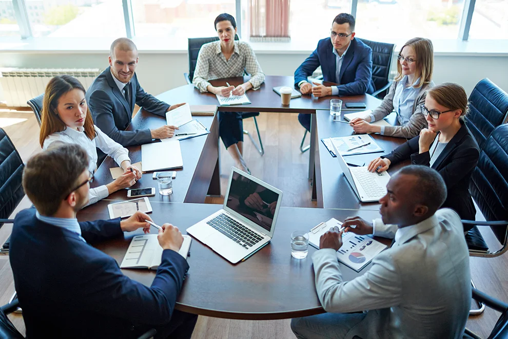 Diverse group of professionals engaged in a meeting around a table with laptops, documents, and discussing business strategies in a modern office.
