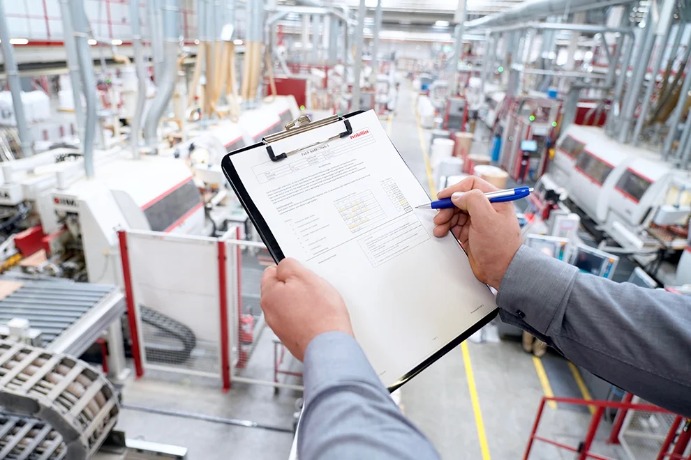 Over-the-shoulder view of a person inspecting a checklist on a clipboard in a modern industrial manufacturing plant with machinery in the background.