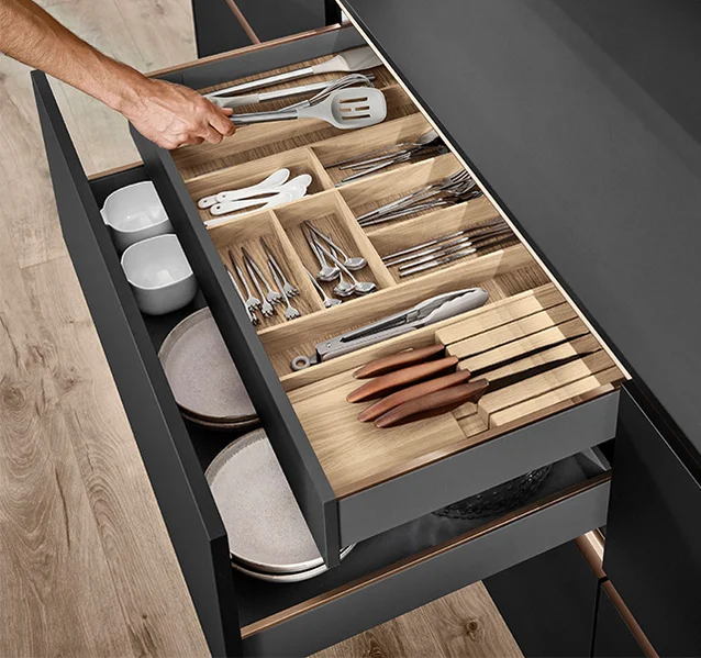 Una mano que se mueve lateralmente coge utensilios de cocina de un organizador de cajón de madera bien ordenado.
