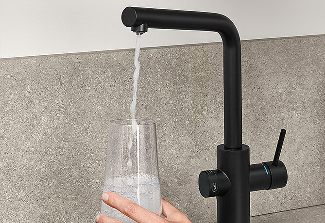 Matte black modern kitchen faucet with a separate filtered water dispenser, filling a clear glass held by a human hand against a gray backsplash.