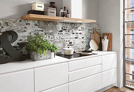 Moderní kuchyňský interiér s bílými skříňkami, kamenným obkladem a dřevěnou poličkou s dekorativními předměty, které vytvářejí útulný a stylový dojem městského domova.