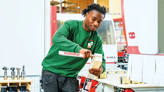 Zwei Auszubildende als Holzmechaniker in der Produktion, die gemeinsam ein Holzstück vermessen und Verarbeitungsschritte planen.