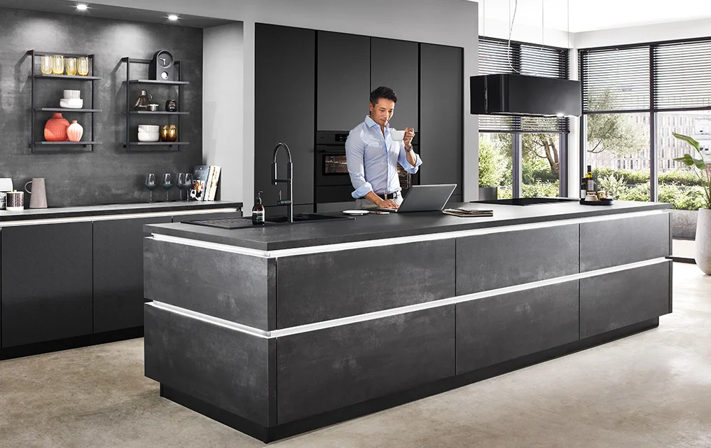 Cucina moderna con frontale LASER 463 in riproduzione di cemento nero, grande isola di cottura con illuminazione integrata, colonne scure e scaffali a giorno con elementi decorativi, uomo che lavora al suo computer portatile.
