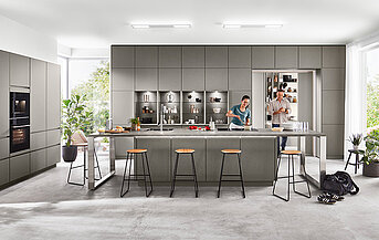Spacious modern kitchen with gray cabinets and central island, bathed in sunlight, where two individuals engage in cooking.