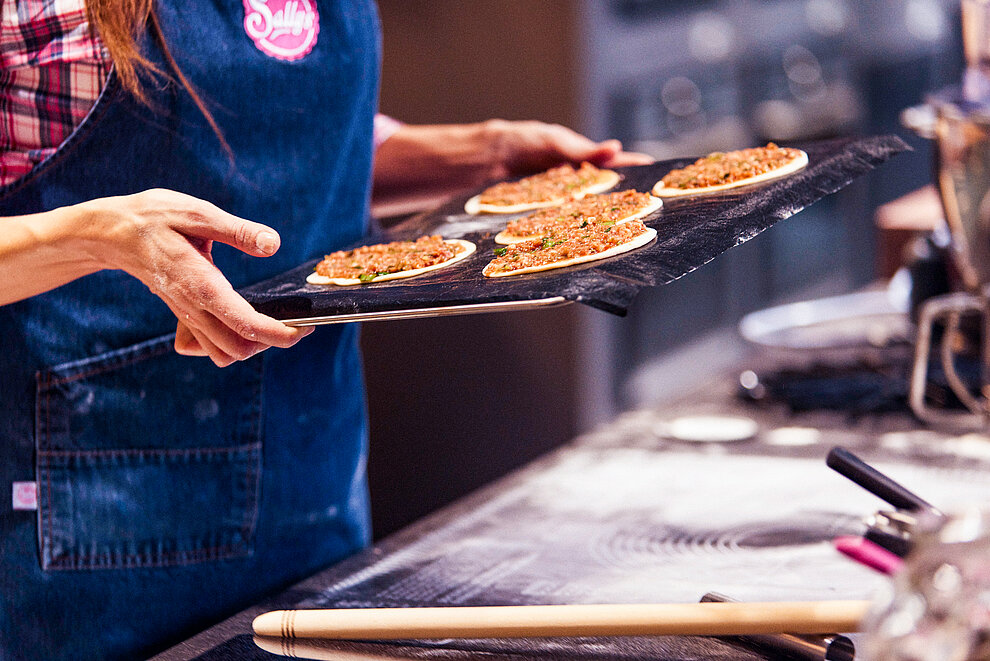 Server in an apron presents a tray of freshly baked, garnished flatbreads in a professional kitchen setting, showcasing culinary expertise and inviting hospitality.