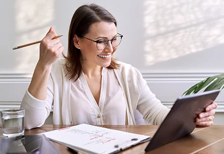 Glimlachende vrouw met bril en pen in de hand, kijkt naar een tablet met een notitieblok naast haar.