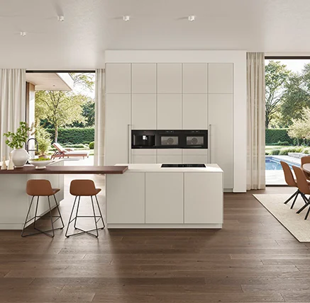 Moderne, helle Küche mit einer zentralen Kochinsel und eleganten Barstühlen an der Theke. Große Fenster eröffnen den Blick auf den Garten.