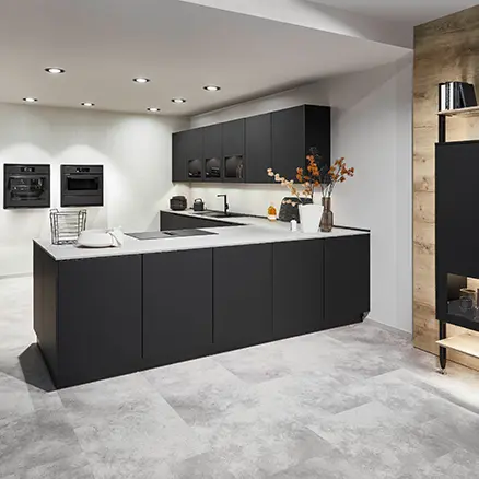Dunkle Küche mit schwarzen Fronten und heller Arbeitsplatte in L-Form, im Hintergrund freistehende Einbaugeräte und ein offenes Regal in Holzoptik.