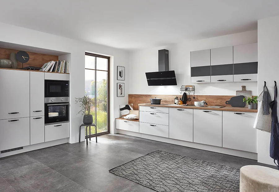 Moderne, helle Küche mit weißen Fronten, Holztheke und großen Fenstern, die Tageslicht hereinlassen.